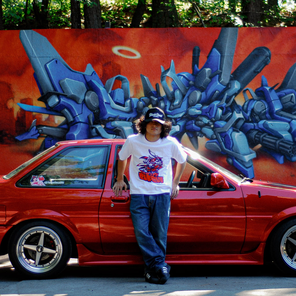 white unisex cotton Kai Oni Dash T-shirt in front of AE 86 with gas guzzler sticker and Mr. Totem graffiti wall