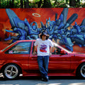 white unisex cotton Kai Oni Dash T-shirt in front of AE 86 with gas guzzler sticker and Mr. Totem graffiti wall